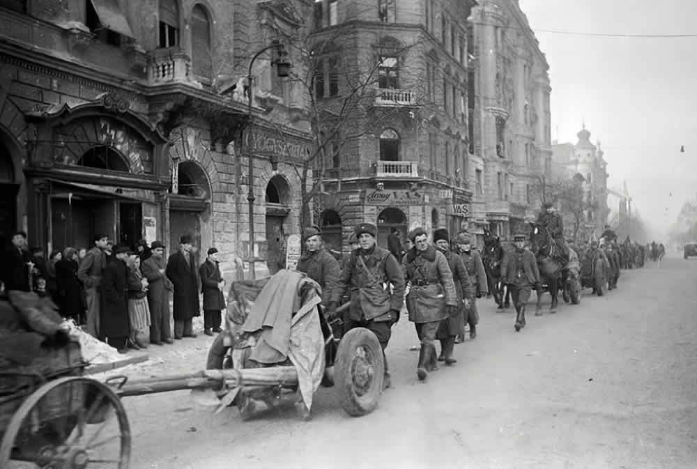 A black-and-white photo of a Soviet infantry column marching down a Boulevard in Budapest with a 45-mm anti-tank gun in the forefront. January 1945.