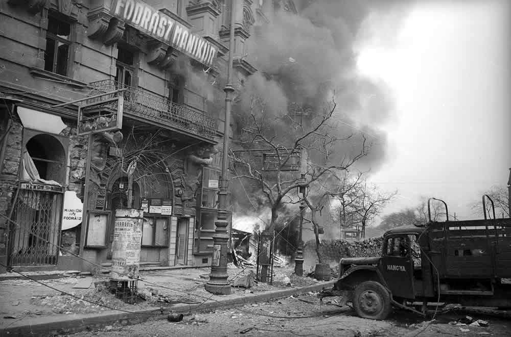 A black-and-white photo showing a burning first floor of a residential building in Budapest, January 1945. There is a damaged truck in front of it.