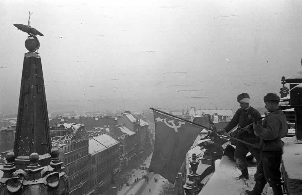 A black-and-white photo showing two Soviet soldiers rising a red flag over the rooftop of a residential building in Budapest, January 1945.